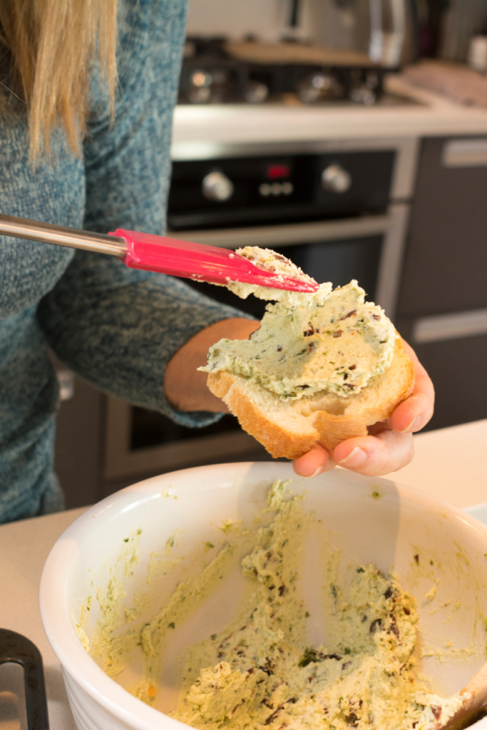 Woman puts cheese mixture on baked bread to make pesto crostini with goat's cheese and sun dried tomatoes